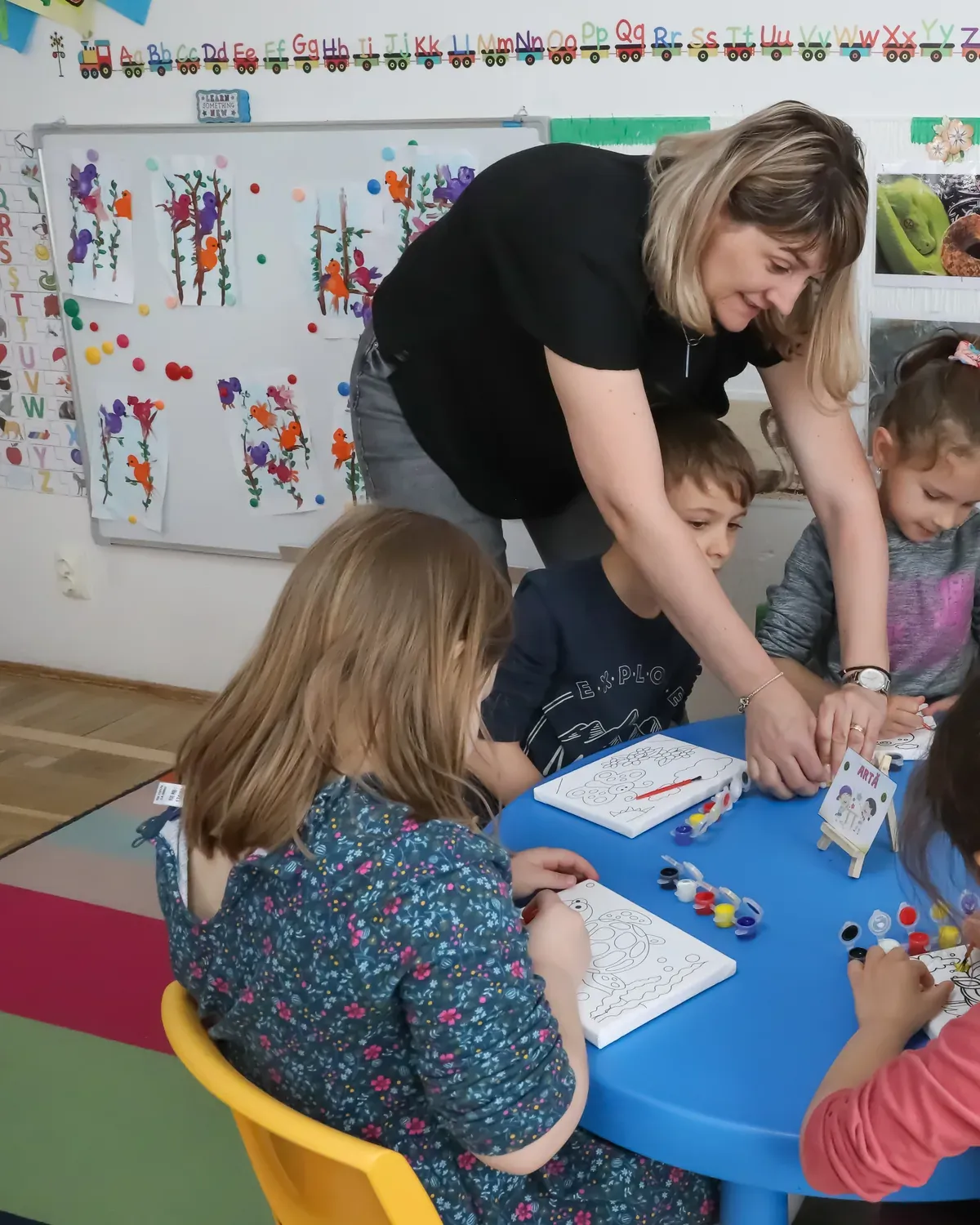 Educatoare aplecată peste masă, lucrând cu copiii grupei